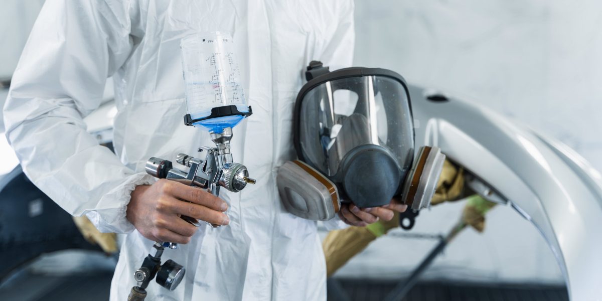 Сlose-up view of car painter with paint gun and protective respirator in hands. Car repair concept. Mechanic in vehicle painting booth